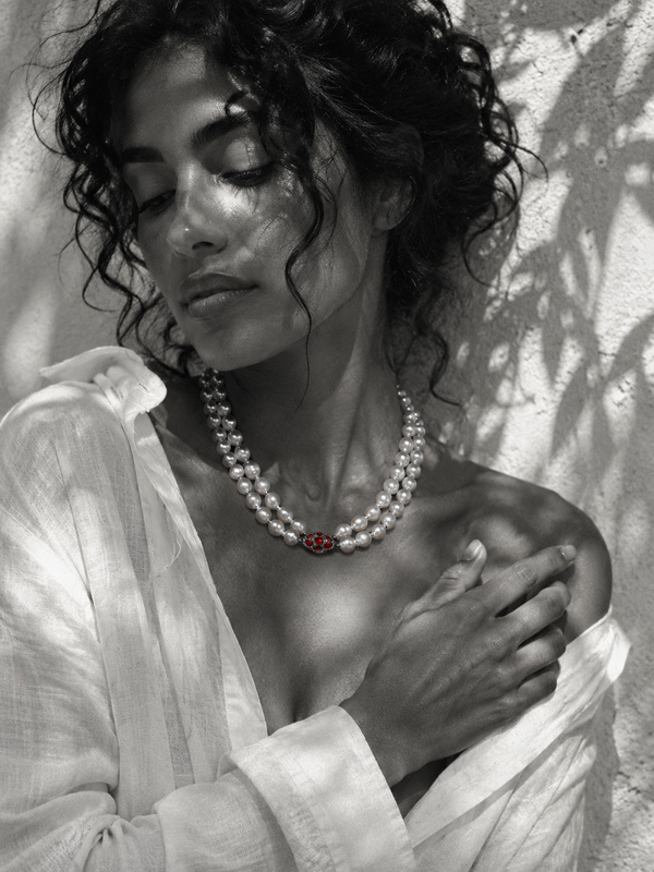 Long baroque pearl necklace doubled at collarbone length, garnet ornament in deep red at center, worn on a model in white linen, Mediterranean light
