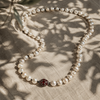 Long baroque pearl necklace laid on natural linen with olive leaf shadows, deep red garnet ornament centered