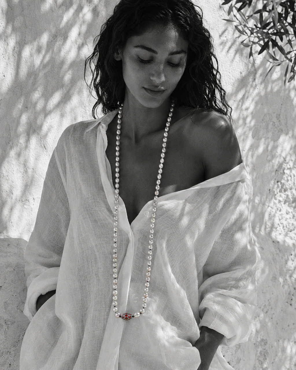 Long baroque pearl necklace worn full length on a model in white linen, Mediterranean light, garnet ornament in deep red