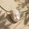 Close-up of baroque freshwater pearl pendant on oxidized dark chain, natural iridescent surface in ivory and pink tones, Mediterranean limestone, leaf shadows