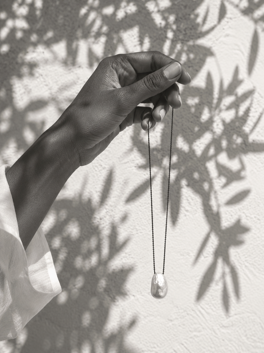 Dark-skinned hand holding a baroque pearl pendant on oxidized chain, whitewashed Mediterranean wall with dappled leaf shadows, white linen sleeve