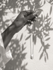 Dark-skinned hand holding a baroque pearl pendant on oxidized chain, whitewashed Mediterranean wall with dappled leaf shadows, white linen sleeve