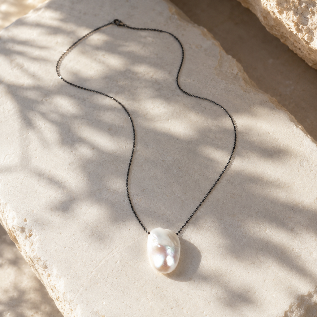 Baroque pearl pendant necklace on oxidized dark chain laid on Mediterranean limestone, olive leaf shadows, natural afternoon light