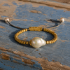 Arabelle bracelet by JLL Pearl — gold-plated square bead clusters and large baroque freshwater pearl on braided cord, resting on weathered blue-painted wood of a Greek fishing boat, warm afternoon light