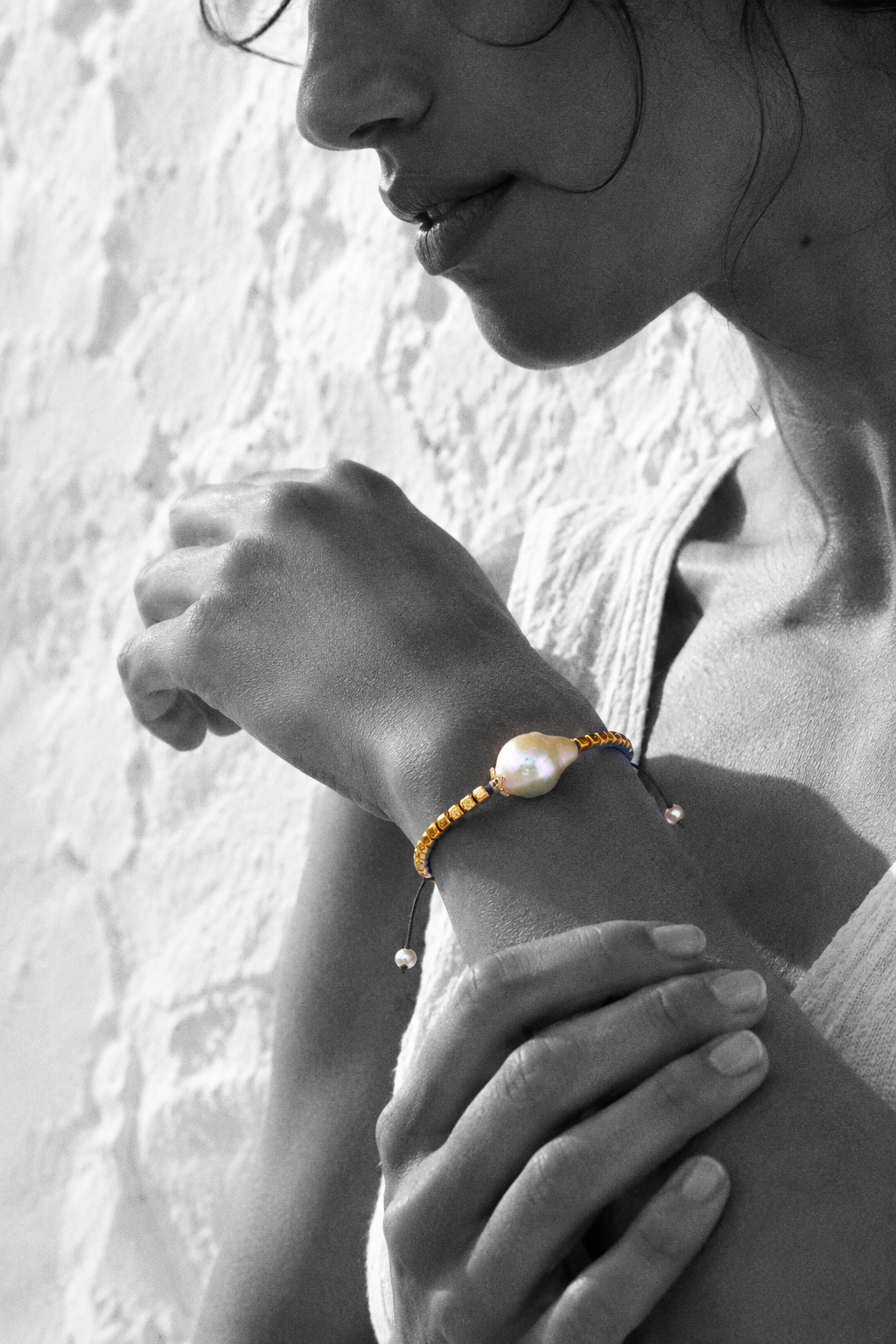 Arabelle bracelet by JLL Pearl — black and white portrait, Mediterranean woman cradling her own wrist, selective colour on gold-plated square beads and baroque freshwater pearl, Greek white wall background
