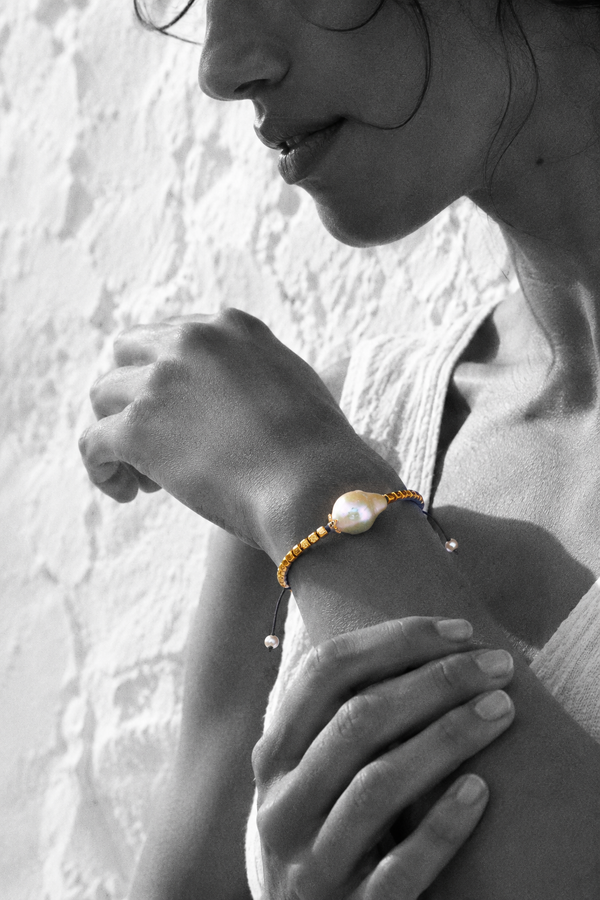 Arabelle bracelet by JLL Pearl — black and white portrait, Mediterranean woman cradling her own wrist, selective colour on gold-plated square beads and baroque freshwater pearl, Greek white wall background
