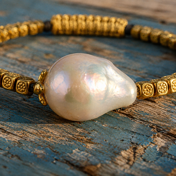 Arabelle bracelet by JLL Pearl — macro close-up on large baroque freshwater pearl with iridescent nacre, flanked by gold-plated engraved square beads with floral motifs, blue-painted wood surface