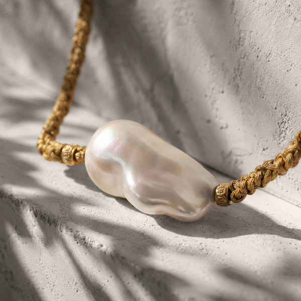 Close-up of Arabelle baroque freshwater pearl on braided gold cord with gold-plated beads, soft iridescence in white, pink and blue tones, olive leaf shadows on whitewashed plaster, handmade by JLL Pearl