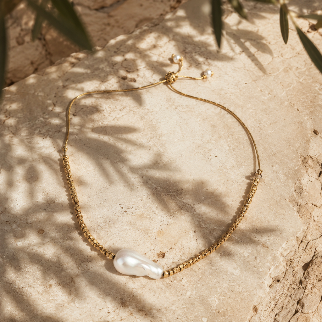 Arabelle baroque pearl gold choker laid on Mediterranean limestone, braided gold cord with gold-plated beads, large baroque freshwater pearl at centre, olive leaf shadows, warm afternoon light, handmade by JLL Pearl