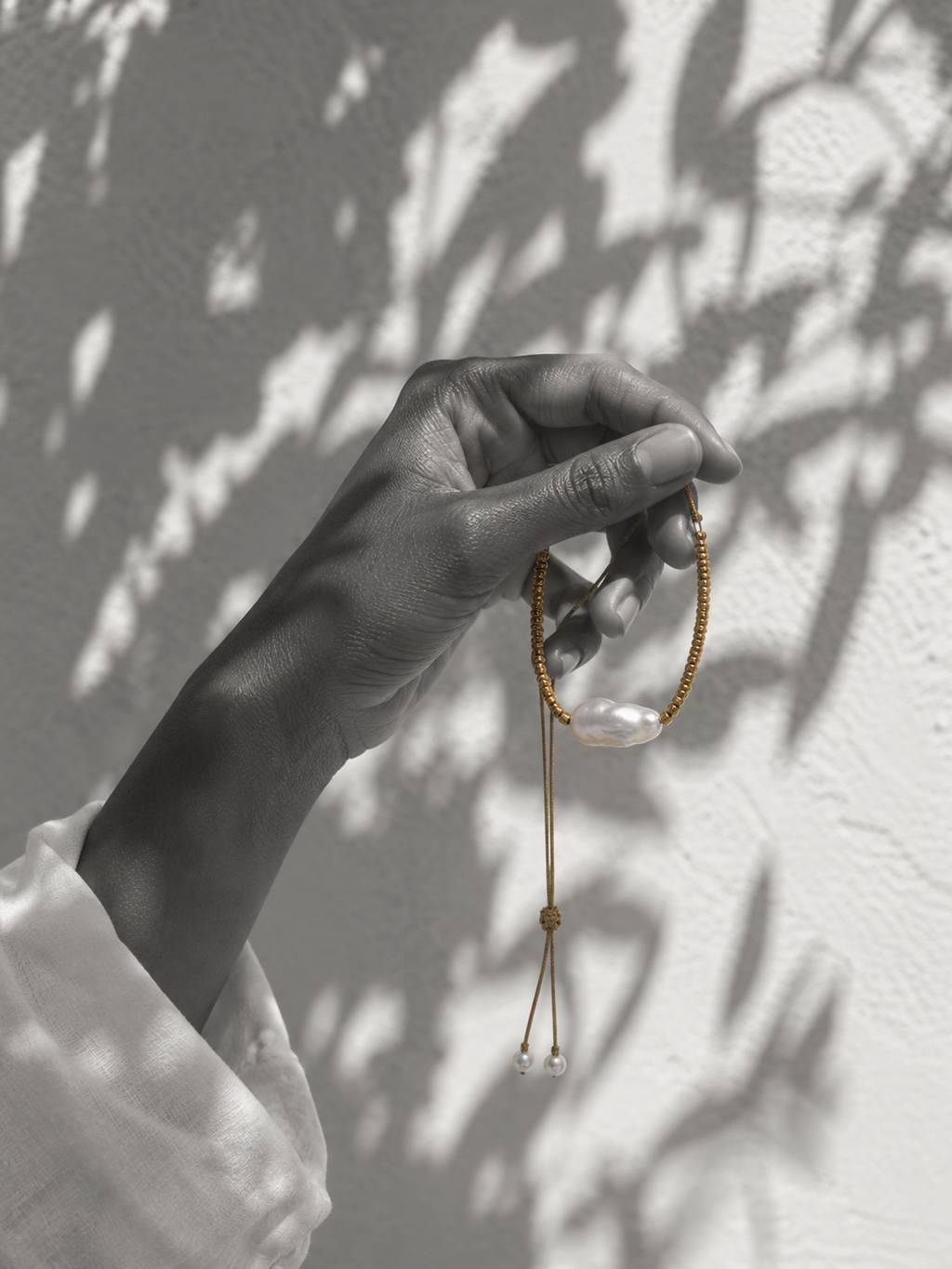 Dark-skinned hand holding Arabelle baroque pearl gold choker by the cord, braided gold with gold-plated beads, baroque freshwater pearl and two small pearl finials, olive branch shadows on whitewashed wall, white linen sleeve, handmade by JLL Pearl
