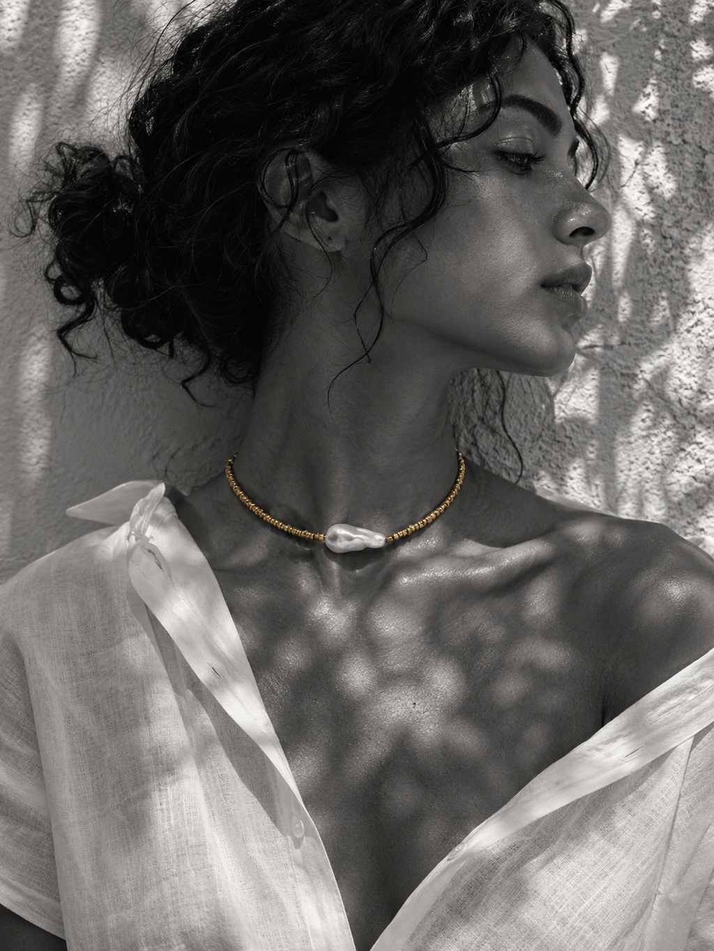 Arabelle baroque pearl gold choker worn at the throat, braided gold cord with gold-plated beads, large baroque freshwater pearl centred, dark curly hair, white linen, olive leaf shadows on whitewashed wall, handmade by JLL Pearl