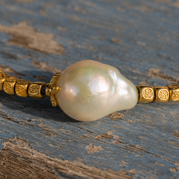 Macro close-up of the Arabelle bracelet — large irregular baroque freshwater pearl with iridescent green and pink nacre, flanked by engraved gold cube beads on a thin black cord, on weathered blue-grey peeling painted wood — JLL Pearl