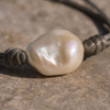 Macro close-up of the Banggai bracelet — large baroque freshwater pearl, cream-white with iridescent rose and green overtones, mounted on a dark oxidized twisted cord with wrapped silver knots, on weathered stone — JLL Pearl
