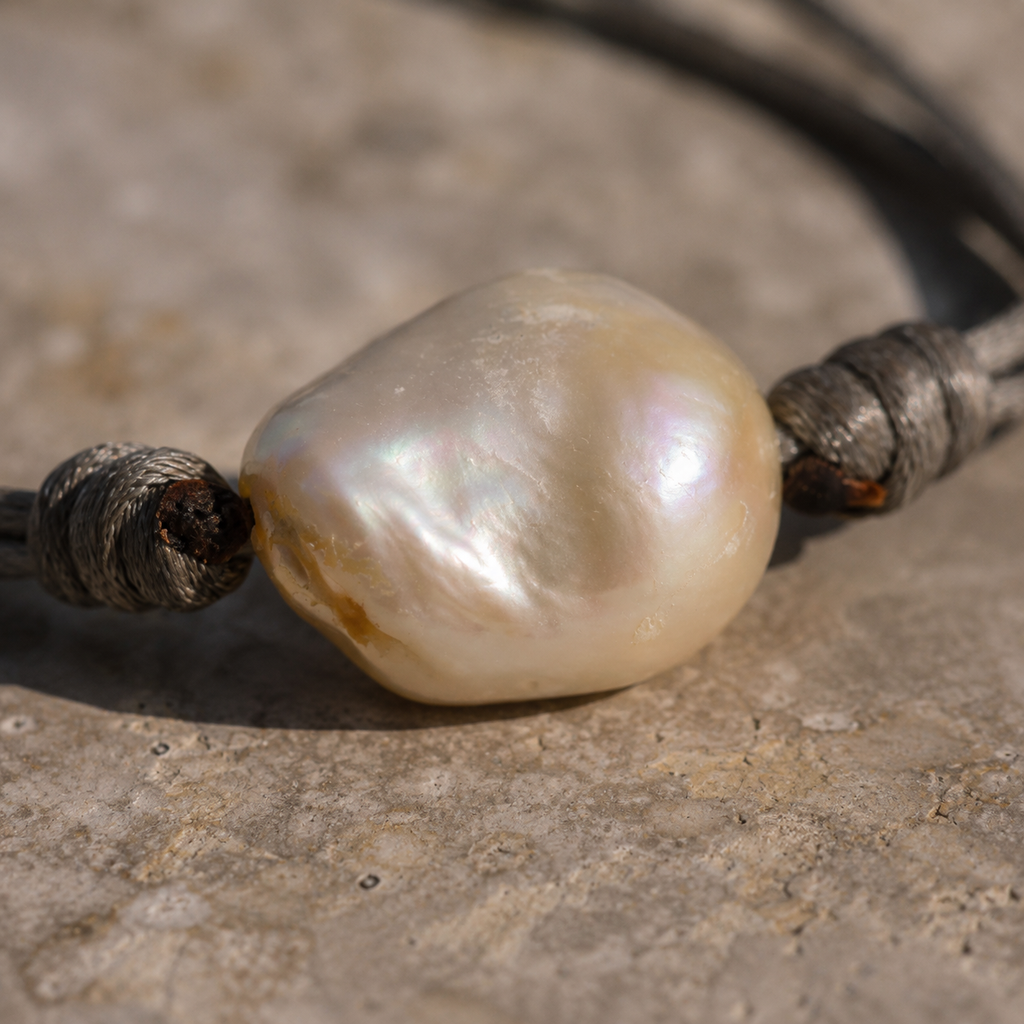 Banggai bracelet by JLL Pearl — macro close-up on large baroque freshwater pearl with iridescent cream-rose-gold nacre, wrapped grey satin knots on each side, grey limestone surface
