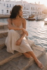 Banggai bracelet by JLL Pearl — Mediterranean woman in sand linen dress seated on ancient stone harbour steps, bracelet with large baroque freshwater pearl visible on wrist resting on knee, Greek port and fishing boats at golden hour