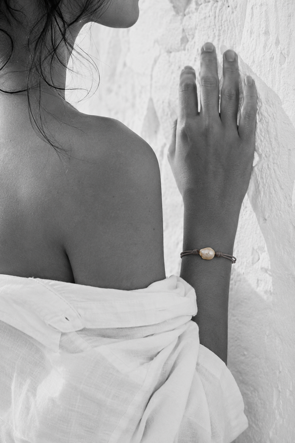 Banggai bracelet by JLL Pearl — black and white portrait, Mediterranean woman, hand flat against rough white Greek wall, selective colour on grey satin cord with single large baroque freshwater pearl