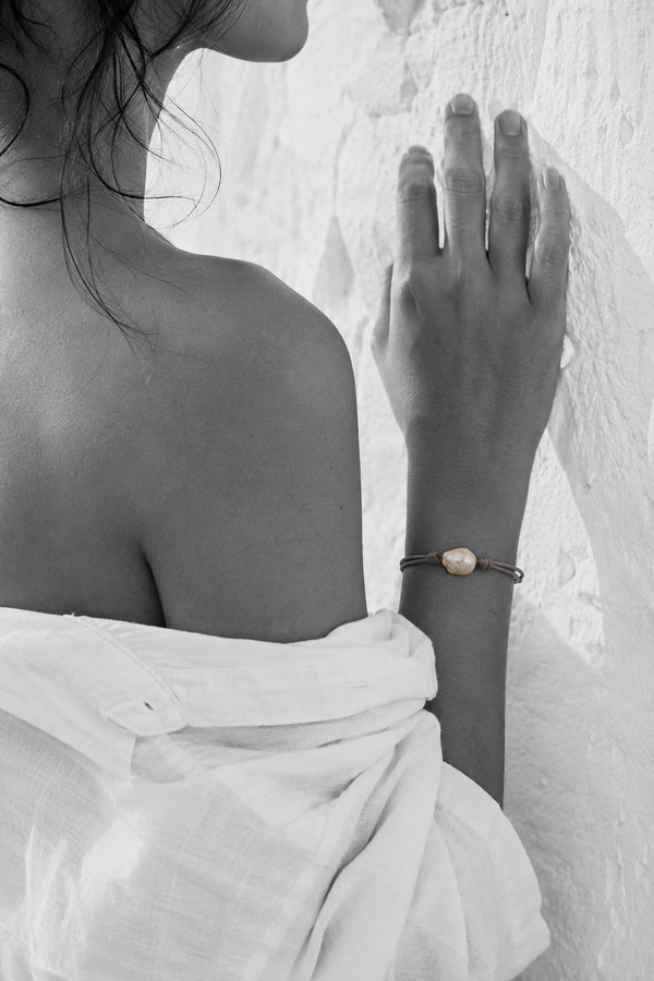Banggai bracelet by JLL Pearl — black and white portrait, Mediterranean woman, hand flat against rough white Greek wall, selective colour on grey satin cord with single large baroque freshwater pearl