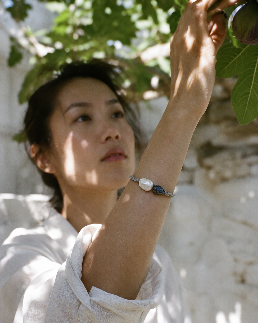 Bocca bracelet by JLL Pearl — East Asian woman in white linen shirt reaching up to pick a fig, grey macramé bracelet with baroque pearl and sapphire teardrop visible on extended forearm, Greek garden
