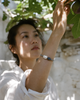 Bocca bracelet by JLL Pearl — East Asian woman in white linen shirt reaching up to pick a fig, grey macramé bracelet with baroque pearl and sapphire teardrop visible on extended forearm, Greek garden
