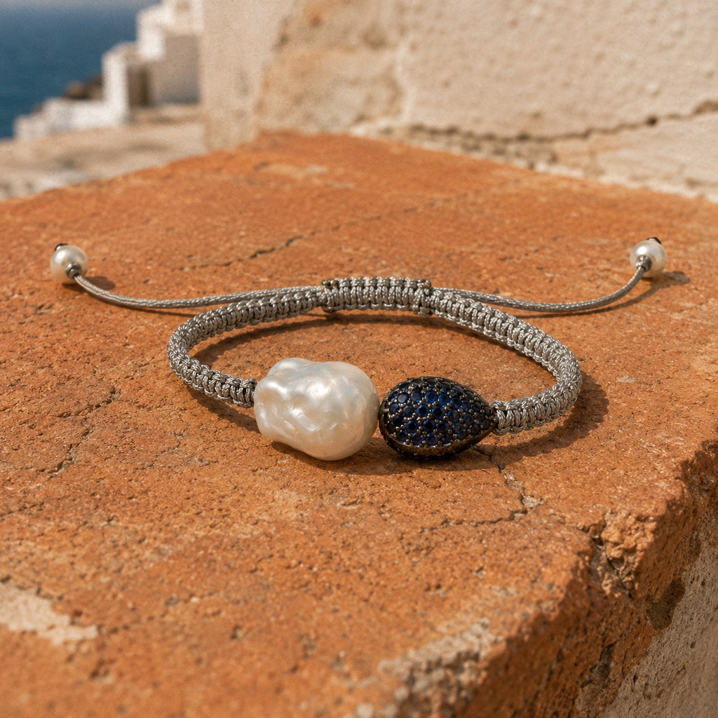 Bocca bracelet by JLL Pearl — grey macramé cord with baroque white freshwater pearl and pavé blue sapphire teardrop, resting on weathered terracotta, Aegean sea in background