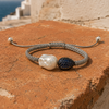Bocca bracelet by JLL Pearl — grey macramé cord with baroque white freshwater pearl and pavé blue sapphire teardrop, resting on weathered terracotta, Aegean sea in background