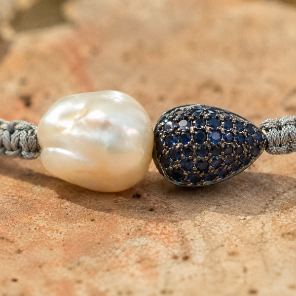 Bocca bracelet by JLL Pearl — macro close-up on baroque white freshwater pearl and navy blue pavé sapphire teardrop side by side on grey macramé cord, warm terracotta surface