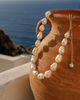 Eden baroque pearl gold flower choker draped over a traditional Greek terracotta amphora on a Mediterranean terrace, deep blue Aegean sea and rocky coastline in the background, warm afternoon light, JLL Pearl
