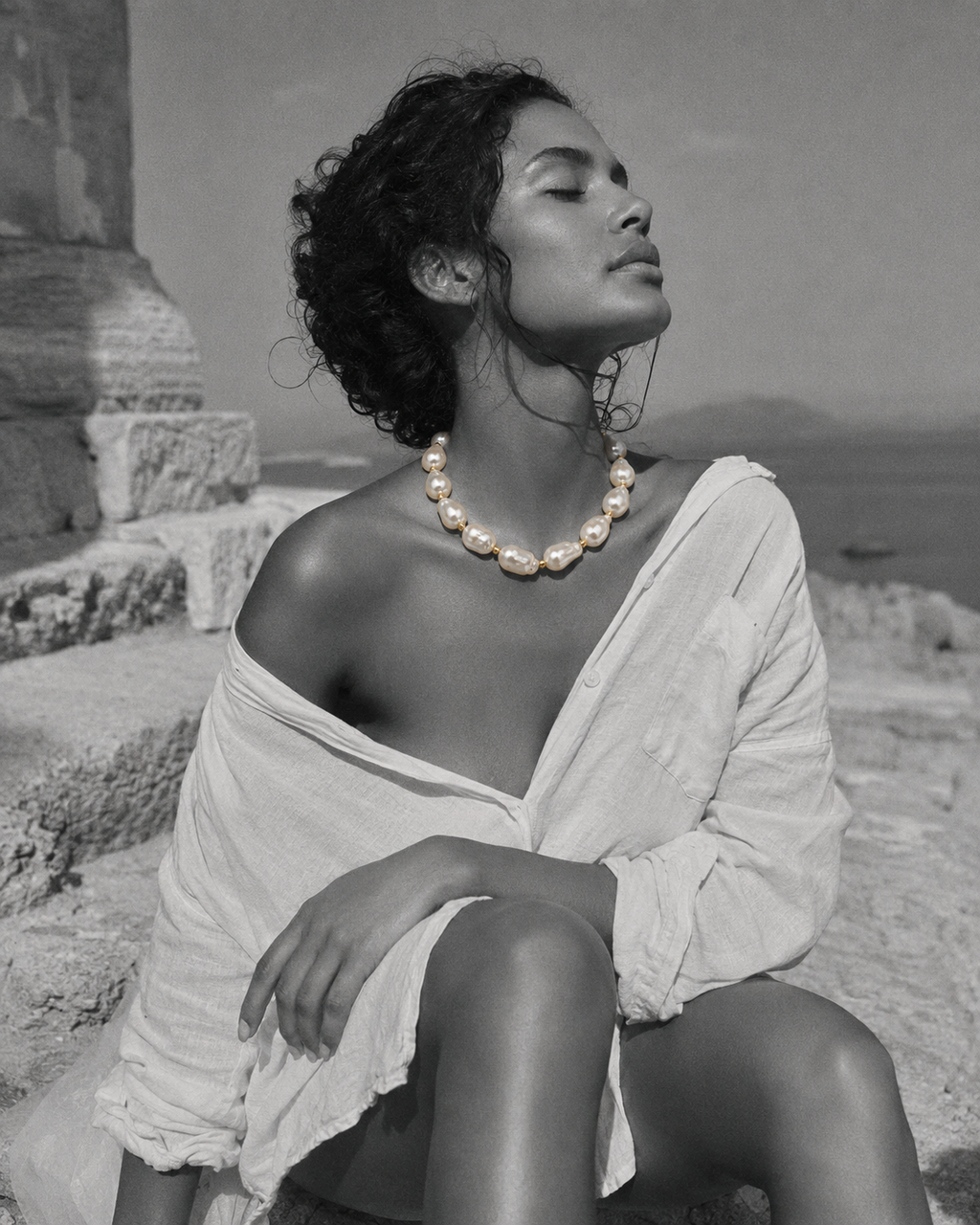 Eden baroque pearl gold flower choker worn against ancient Greek ruins, woman seated on stone steps with eyes closed facing the Aegean sea, selective colour necklace against black and white scene, handmade by JLL Pearl