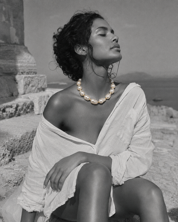 Eden baroque pearl gold flower choker worn against ancient Greek ruins, woman seated on stone steps with eyes closed facing the Aegean sea, selective colour necklace against black and white scene, handmade by JLL Pearl