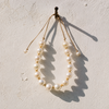 Ella round pearl gold flower choker hanging from iron nail on whitewashed Greek wall casting sharp shadow in Mediterranean light