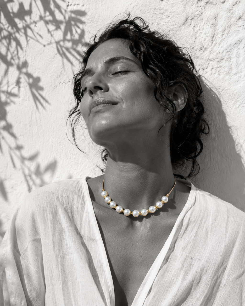Ella round pearl gold flower choker worn at the throat, woman head tilted back eyes closed in Mediterranean sun against whitewashed wall
