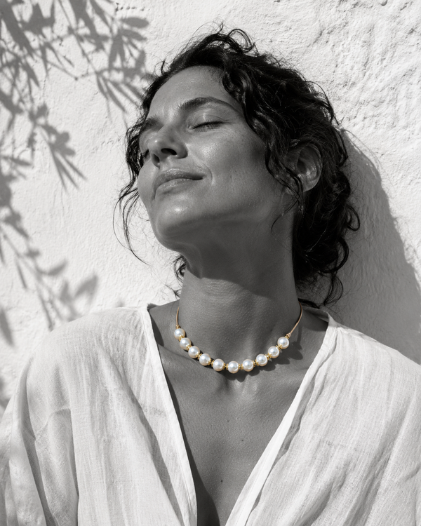 Ella round pearl gold flower choker worn at the throat, woman head tilted back eyes closed in Mediterranean sun against whitewashed wall