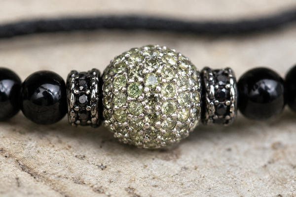 Hydra black onyx peridot necklace detail, central silver sphere pavé-set with pale green peridot flanked by black spinel toupies and onyx beads, macro on pale sandstone, handmade by JLL Pearl