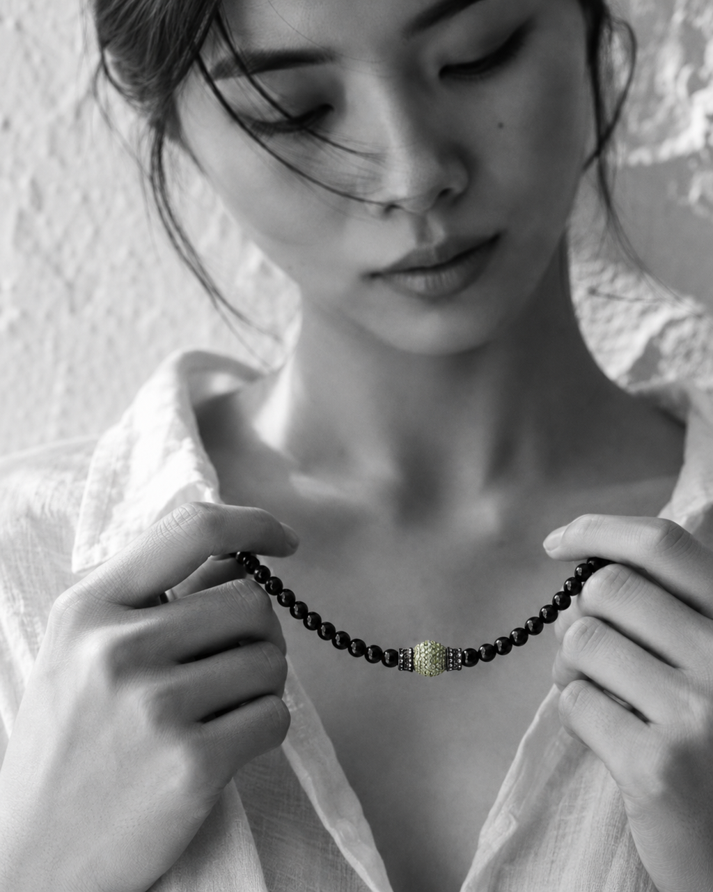 Hydra black onyx peridot necklace held in both hands by Asian model, face softly out of focus looking down, peridot sphere in natural green, whitewashed wall, handmade by JLL Pearl