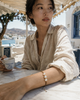 Young woman wearing the Ios gold macramé pearl bracelet, forearm resting on a marble café table, holding a Greek coffee cup, sea view and whitewashed buildings in the background, natural Mediterranean light