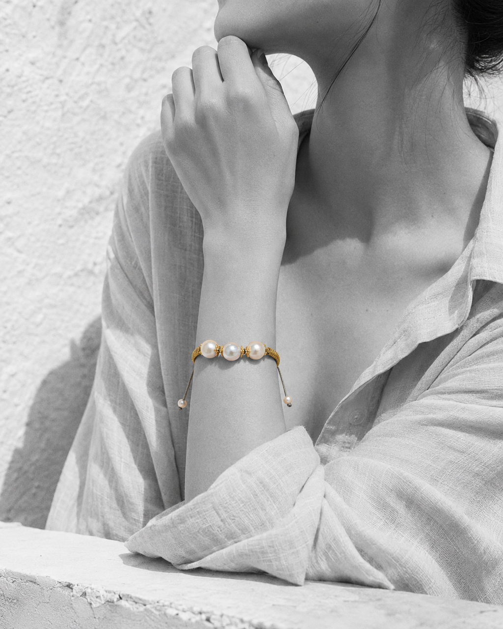 Woman wearing the Ios gold macramé pearl bracelet — three round freshwater pearls with gold flower spacers on a golden satin cord — portrait in black and white with selective colour on the bracelet, whitewashed Greek wall background