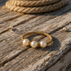Ios gold macramé pearl bracelet with three round freshwater pearls and gold flower spacers, resting on sun-bleached weathered wood planks, coiled rope in background, warm Mediterranean afternoon light