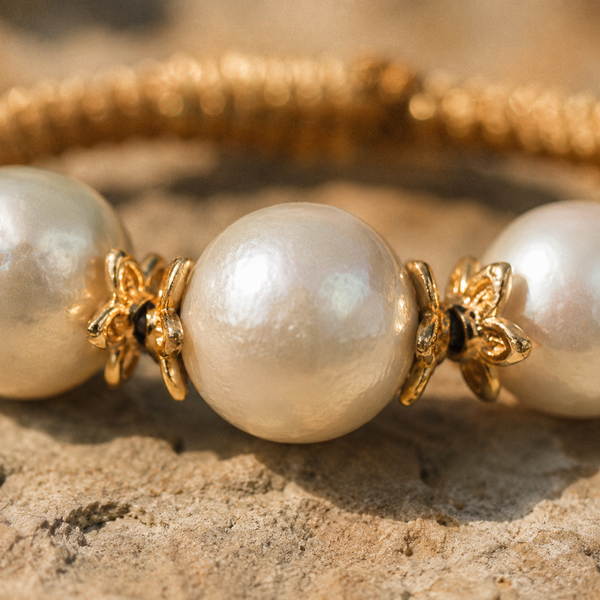 Macro close-up of the Ios bracelet — central round freshwater pearl with small gold flower-shaped bead cap spacers on each side, golden macramé cord visible above, warm raking light on natural stone surface