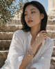 Karla bracelet worn on the wrist of a young woman sitting on ancient Greek stone steps — bronze macramé cord, amethyst and aquamarine marquise stone cluster in rose gold, pink baroque pearl pendant, olive tree and Greek temple in the background — JLL Pearl