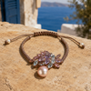 Karla bracelet — bronze macramé cord, asymmetric cluster of pale amethyst and aquamarine marquise-cut stones in rose gold settings, pink baroque freshwater pearl pendant, resting on warm sandstone with blue Greek shutters and Aegean sea in the background — JLL Pearl