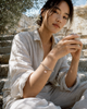 Karla bracelet by JLL Pearl — East Asian woman seated on ancient Greek stone steps, wearing sandy macramé bracelet with raw crystal cluster and baroque pearl, warm summer light