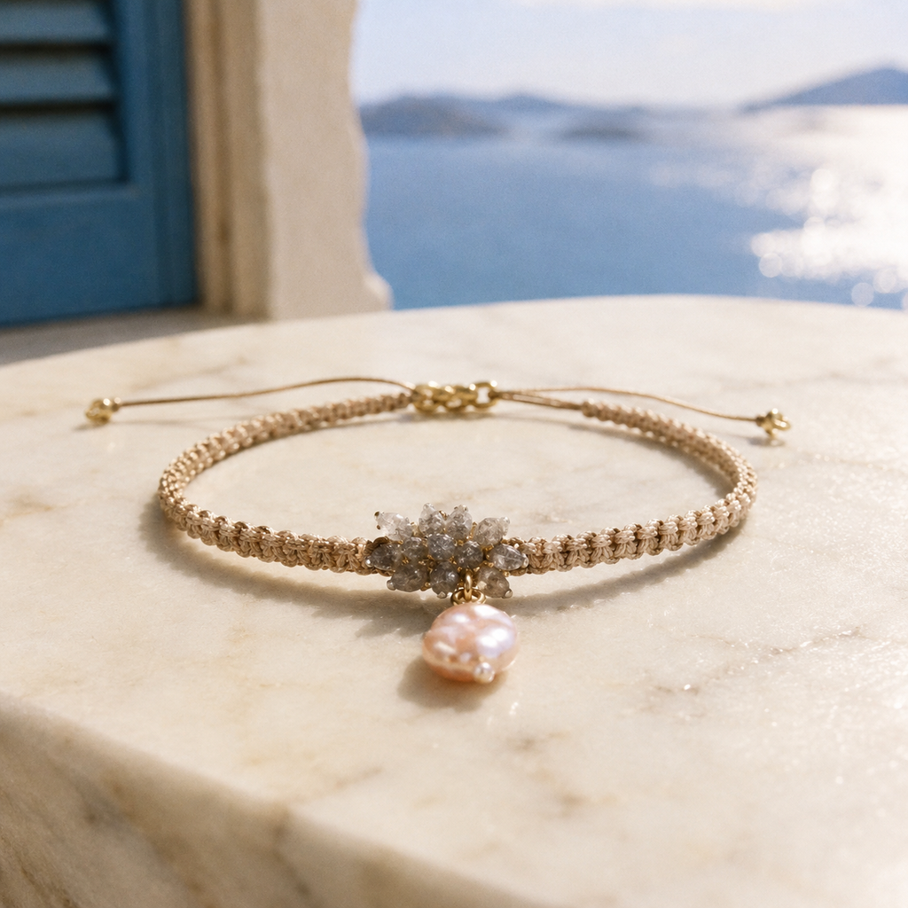 Karla bracelet by JLL Pearl — sandy beige macramé cord with raw grey crystal cluster and baroque pink freshwater pearl, resting on white marble, Aegean sea view in background
