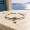 Karla bracelet by JLL Pearl — sandy beige macramé cord with raw grey crystal cluster and baroque pink freshwater pearl, resting on white marble, Aegean sea view in background