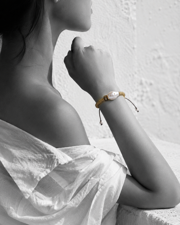 Kea baroque pearl gold macramé bracelet on wrist raised near face, black and white portrait with selective colour on bracelet