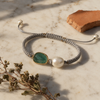 Killi handmade macramé bracelet flat lay — emerald and freshwater pearl on grey knotted cord, marble surface with terracotta shard and dried herbs, Greece
