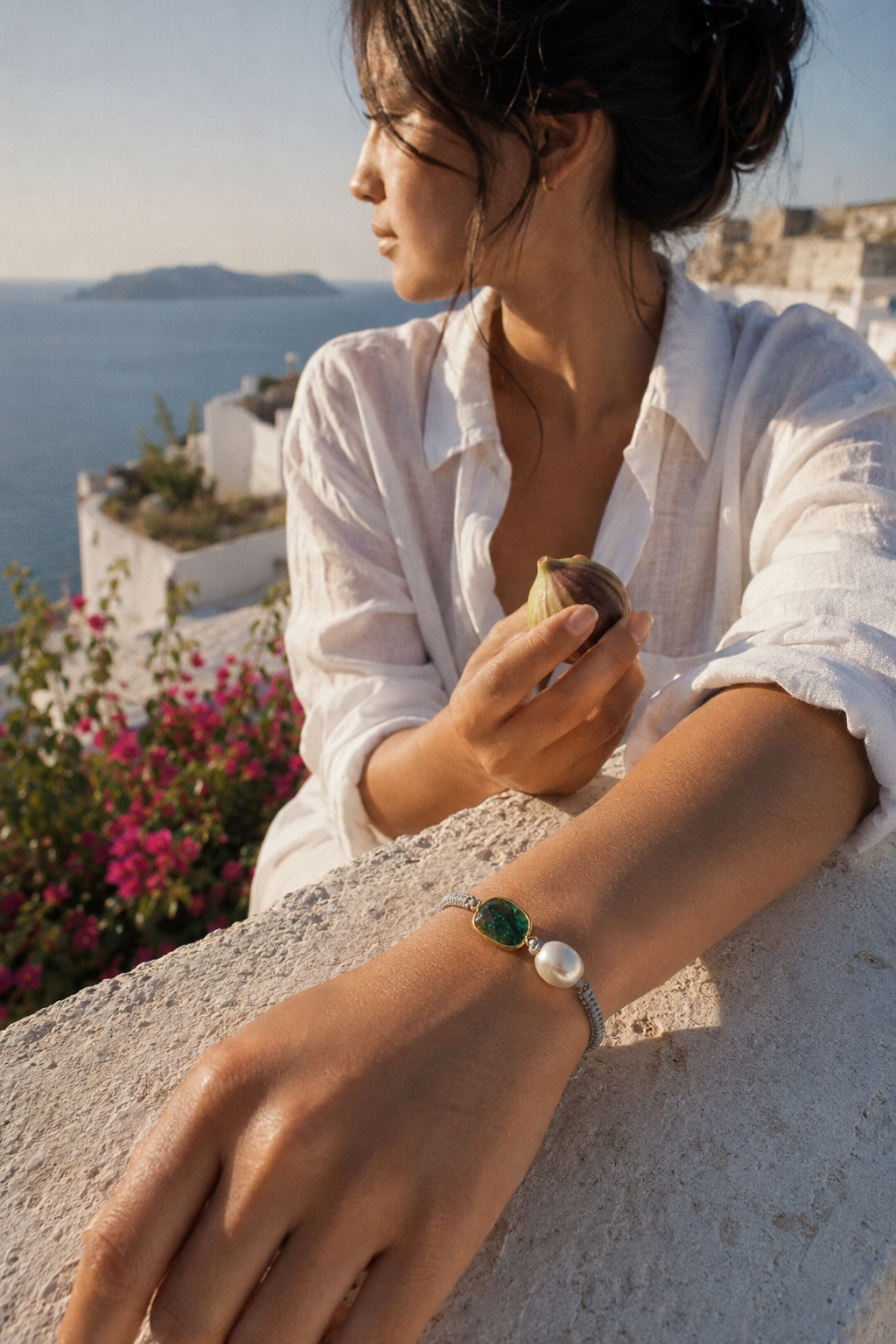 Asian woman wearing Killi emerald and pearl macramé bracelet on whitewashed Greek terrace, white linen shirt, Aegean sea and bougainvillea in background