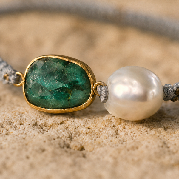 Close-up of Killi bracelet central element — raw emerald in antique gold bezel beside baroque freshwater pearl on grey macramé cord, natural sand surface