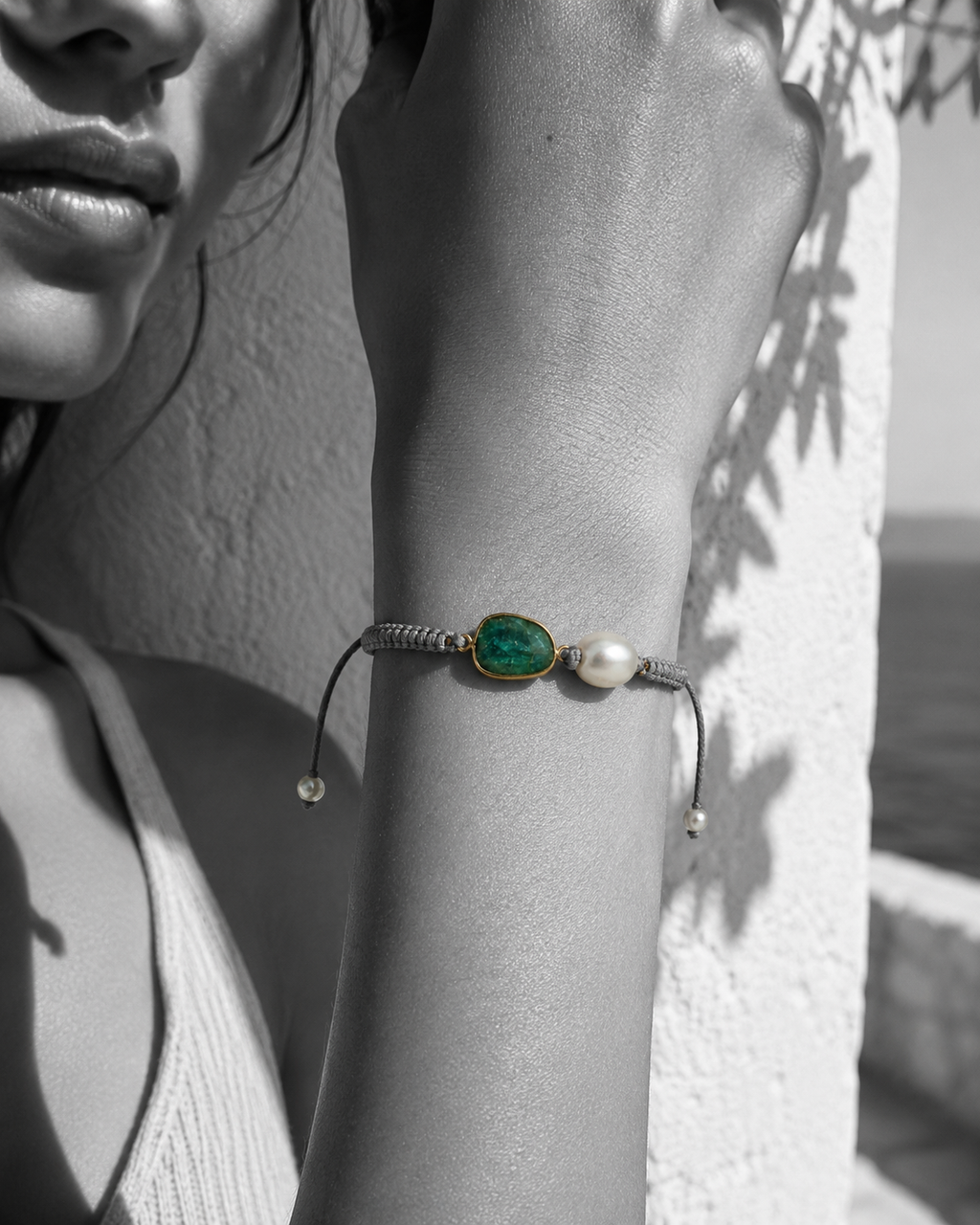 Killi bracelet worn on wrist — raw emerald in gold bezel and baroque freshwater pearl on grey macramé cord, black and white photograph with bracelet in color, Greek white wall background