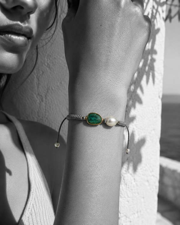 Killi bracelet worn on wrist — raw emerald in gold bezel and baroque freshwater pearl on grey macramé cord, black and white photograph with bracelet in color, Greek white wall background