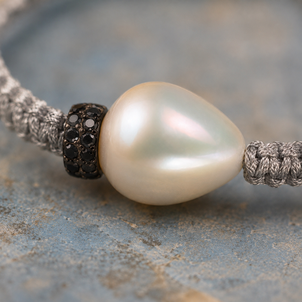 Macro close-up of the Killini bracelet — large baroque freshwater pearl with iridescent nacre, two stacked red sapphire pavé rondelles at its base, light grey macramé cord on both sides, on weathered blue-grey painted stone — JLL Pearl